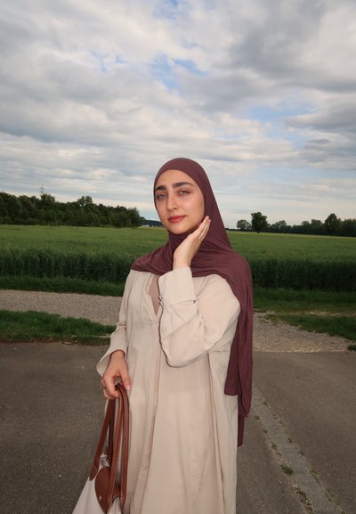 Vestido largo de manga larga color beige claro con corte holgado, combinado con un hiyab marrón oscuro. Sosteniendo un bolso marrón, de pie frente a un fondo de campo verde.