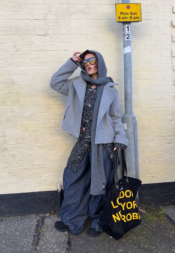 Person in layered gray clothing and oversized pants leans against a wall, holding a black tote bag with yellow text near a street sign.