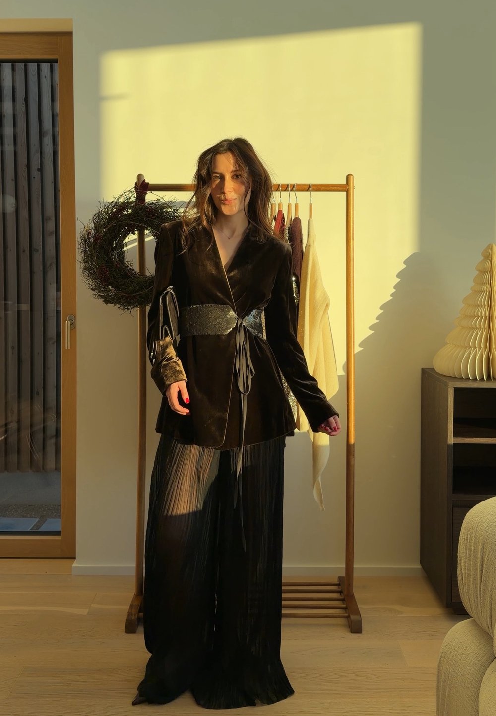 Femme en tenue de velours noir posant à l'intérieur près d'un portant en bois avec des vêtements accrochés et une couronne décorative, baignée de lumière chaude du soleil.