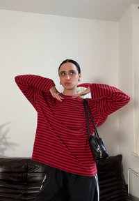 Chemise oversize rayée rouge et noire avec manches larges, associée à un pantalon noir. Petit sac à main noir avec détails zippés. Fond clair.