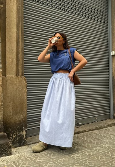 Camiseta azul corta, falda larga azul clara con cintura elástica y zuecos beige. De pie contra un fondo de ondulado gris, sosteniendo una taza.