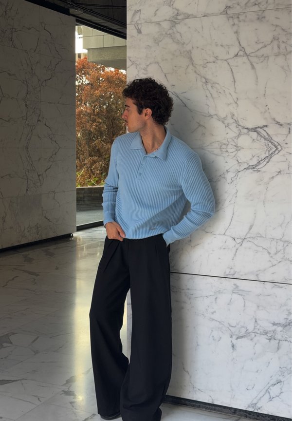 Hombre con cabello rizado que lleva un suéter azul claro acanalado y pantalones anchos negros se apoya contra una pared blanca de mármol, mirando hacia su izquierda.