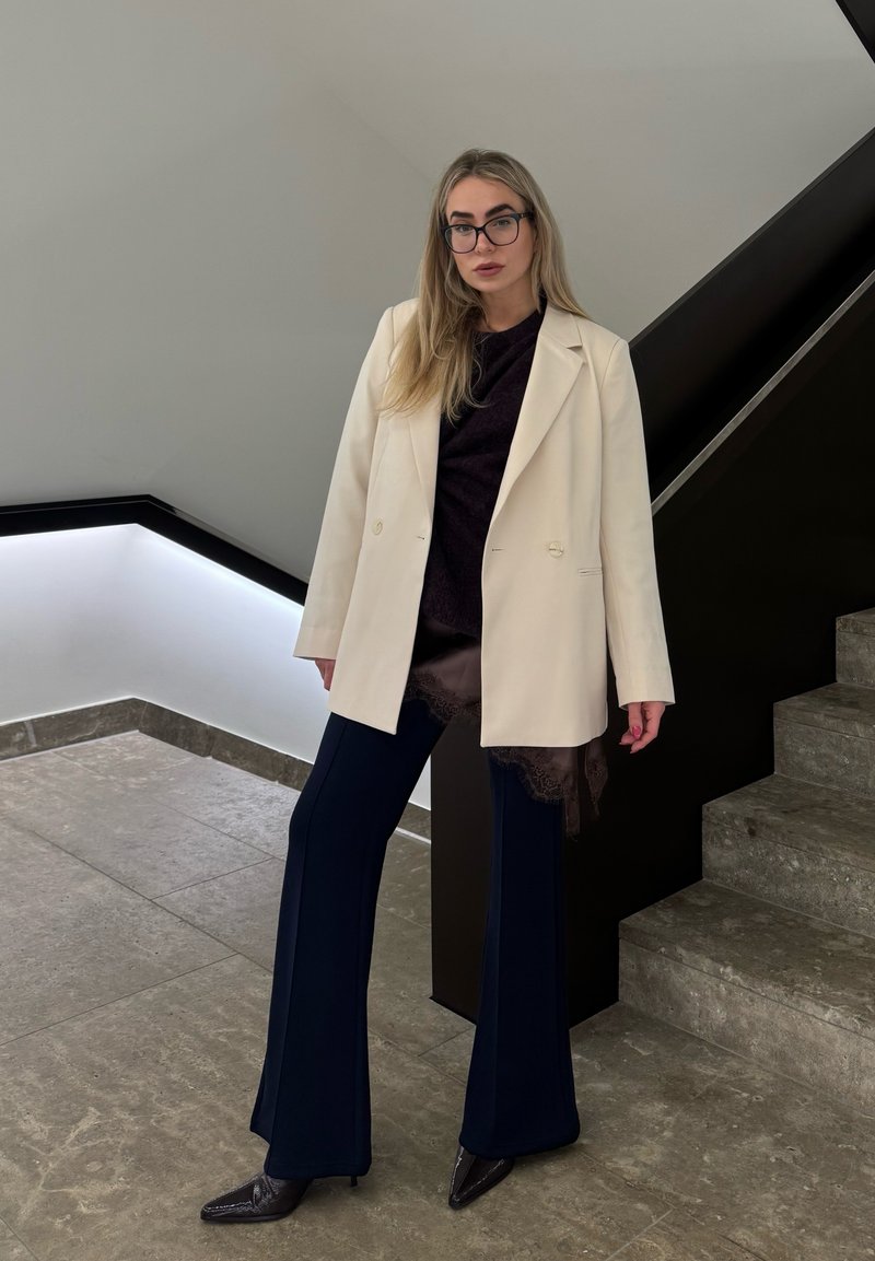 Femme portant des lunettes, un blazer blanc, un haut foncé avec un ourlet en dentelle, un pantalon évasé bleu marine et des chaussures pointues, debout près des escaliers à l'intérieur.