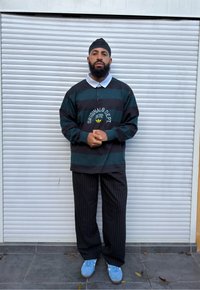 Homme barbu portant une chemise à manches longues rayée noir et vert foncé, un pantalon noir à fines rayures, des baskets bleues et un couvre-chef noir, debout en plein air.