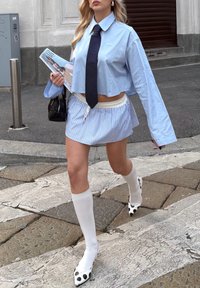Kvinde går på en brostensbelagt gade iført en kort blå skjorte, sort slips, stribet mininederdel, hvide knæstrømper og sko med prikker, bærende på en taske og et magasin.
