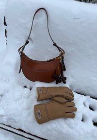 Sac à main en cuir marron avec des breloques en forme de clé et de chien suspendues à un banc couvert de neige, gants UGG couleur fauve reposant sur la neige en dessous.