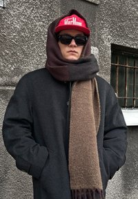 Črn plašč z rjavim, teksturiranim šalom, pritrjenim okoli vratu. Rdeča kapa z belim napisom. Temna sončna očala. Siva stena v ozadju.