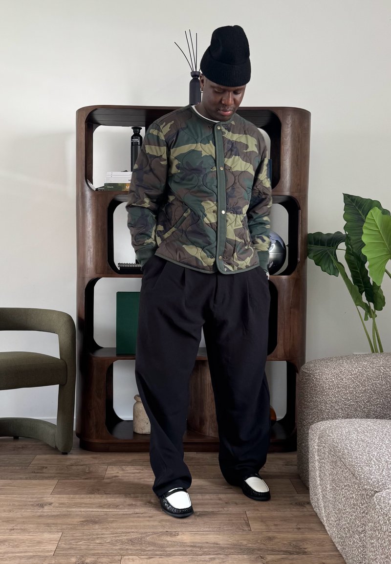 Homme portant un bonnet noir, une veste matelassée camouflage et un pantalon large noir, debout dans un salon moderne avec une étagère en bois et des plantes vertes.