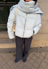 Personne en doudoune bleu clair, pantalon large rayé, gants gris, chaussures bleues, portant un sac à main beige, debout sur un trottoir carrelé à côté d'un mur.