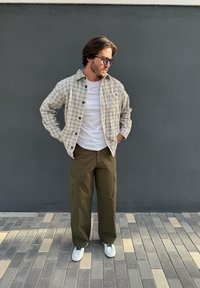 Chemise à carreaux beige et noire boutonnée par-dessus un T-shirt blanc. Pantalon cargo vert olive avec poches latérales. Baskets blanches avec des accents verts.