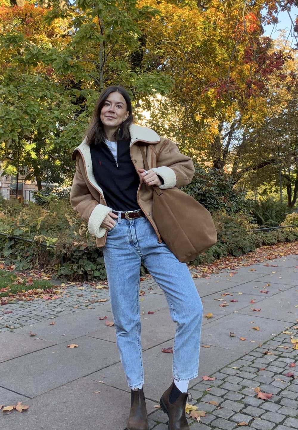 Braune Schaffelljacke, schwarzes Zip-Top, hellblaue Jeans, braune Stiefeletten. Tan Tasche, draußen mit buntem Herbstlaub im Hintergrund.