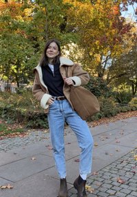Braune Schaffelljacke, schwarzes Zip-Top, hellblaue Jeans, braune Stiefeletten. Tan Tasche, draußen mit buntem Herbstlaub im Hintergrund.