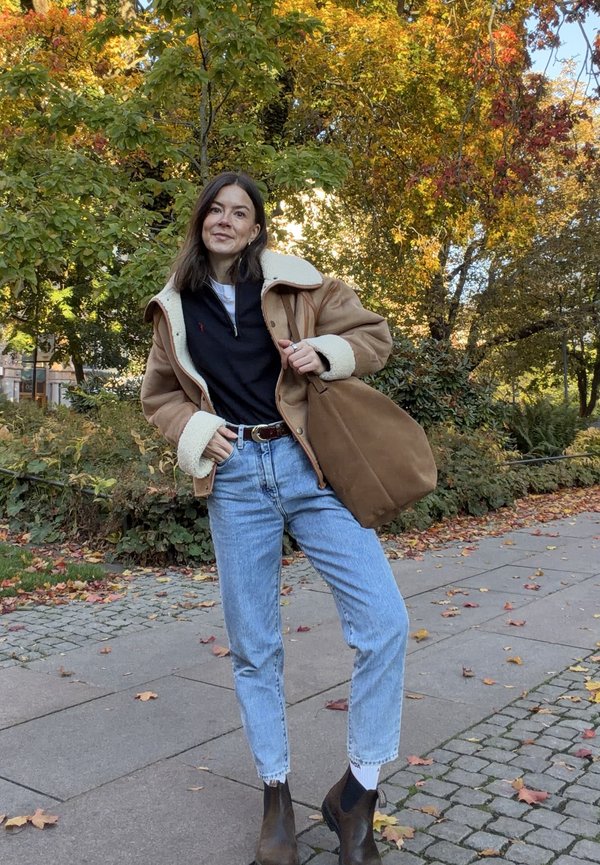 Brun shearlingjakke, sort lynlåstrøje, lyseblå jeans, brune ankelstøvler. Tan taske, udendørs med farverigt efterårsblade i baggrunden.