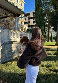 Femme en veste de fourrure sombre tenant un petit chien brun en pull à motifs, debout dans un jardin ensoleillé avec des bâtiments historiques et modernes en arrière-plan.