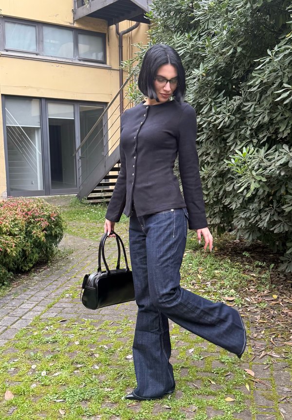 Donna con taglio capelli nero a caschetto, indossa una giacca nera abbottonata e jeans svasati, tiene una borsa nera, posa all'aperto vicino al verde.