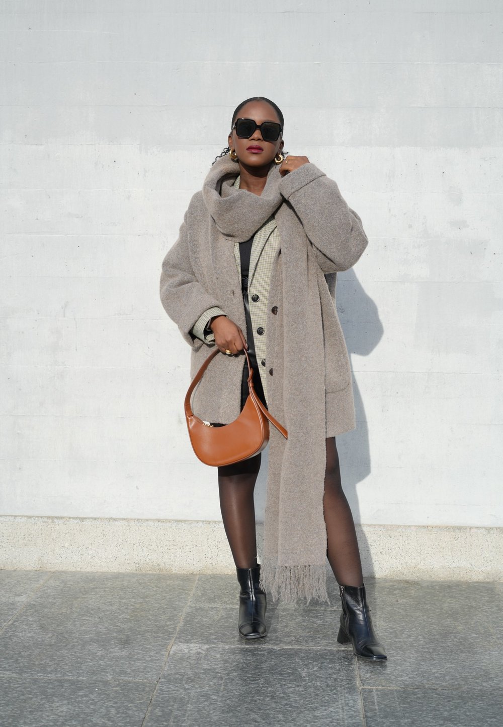 Hallikas ülerõivas koos sobiva salliga, kihtidena kantud kontrollitud bleiseri kohal. Mustad saapad ja pruun õlakott lõpetavad välimuse.