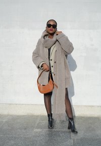 Hallikas ülerõivas koos sobiva salliga, kihtidena kantud kontrollitud bleiseri kohal. Mustad saapad ja pruun õlakott lõpetavad välimuse.