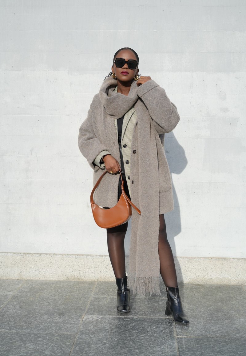 Γκρι υπερμεγέθη παλτό με ταιριαστό κασκόλ, επενδεδυμένο με ένα καρό σακάκι. Μαύρες μπότες και μια καφέ τσάντα ώμου συμπληρώνουν την εμφάνιση.