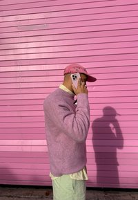 Rozā džemperis, gaiši zaļas bikses un rozā cepure; tur baltu viedtālruni pret rozā sienu ar horizontālām līnijām. Redzama ēna.