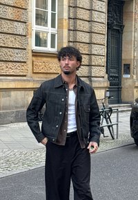 Giovane con capelli ricci indossa una giacca scura, una camicia a quadri, una canotta bianca e pantaloni neri, mentre si trova su un marciapiede cittadino vicino a una bicicletta.