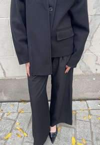 Persoon in een zwart op maat gemaakt jasje, wijde broek en puntige zwarte schoenen die op een met bladeren bedekt stenen trottoir staat tegen een stenen muur.