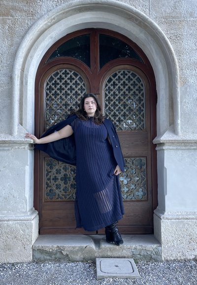 Vestido azul marino con diseño texturizado, combinado con un abrigo oscuro. De pie frente a una puerta de madera ornamentada con un patrón enrejado. Botas negras.