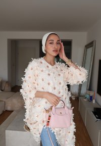 Tunic roz deschis cu un model floral, cu petale texturate și stratificate, accesorizată cu o geantă mică roz, decorată cu mărgele colorate și accente aurii.