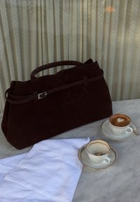 Mørkebrun semsket skinnveske, hvit klut, og to vintage hvite kaffekopper med tallerkener og skjeer på et marmorbord.