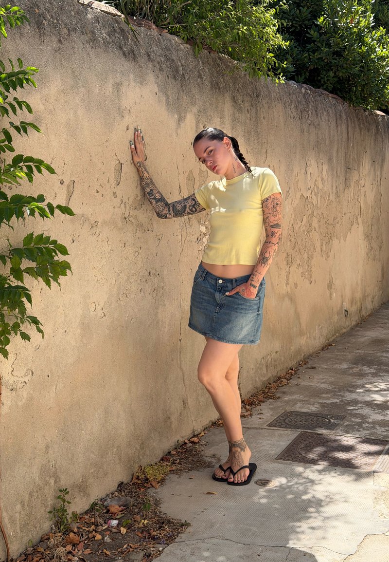 Jeune femme tatouée portant un t-shirt jaune, une jupe en jean et des sandales, s'appuie contre un mur beige texturé à l'extérieur par temps ensoleillé.