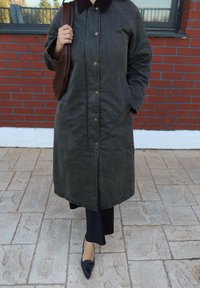 Persona con un lungo cappotto scuro, pantaloni neri e scarpe scure a punta, che tiene una borsa marrone a tracolla, in piedi su un marciapiede piastrellato accanto a un muro di mattoni.