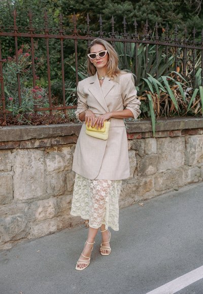 Blazer beige de lino sobre un vestido de encaje transparente, sosteniendo un bolso amarillo. Sandalias nude con tiras, gafas de sol y un collar llamativo visibles.