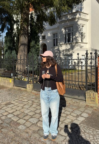 Mujer con gorra rosa, cárdigan negro y vaqueros azul claro, de pie sobre un camino de adoquines junto a una verja de hierro frente a un edificio blanco.