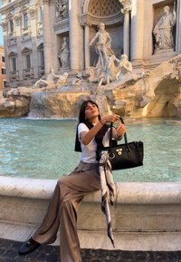 Mustast nahast käekott hõbedaste detailidega, valge t-särk, pruunid laiad püksid ja mustriline siidmütsid kivist purskkaevu taustal.