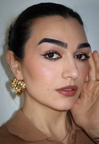 Goldene herzförmige Ohrringe mit strukturierten Details. Hellbraunes Oberteil mit Kragen. Glatte Haut, neutrales Make-up und dunkel, leicht welliges Haar.