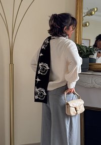 Pull blanc texturé, pantalon gris à jambes larges, écharpe noire avec motif de crâne et sac à main crème avec fermoir. Réflexion en miroir en arrière-plan.