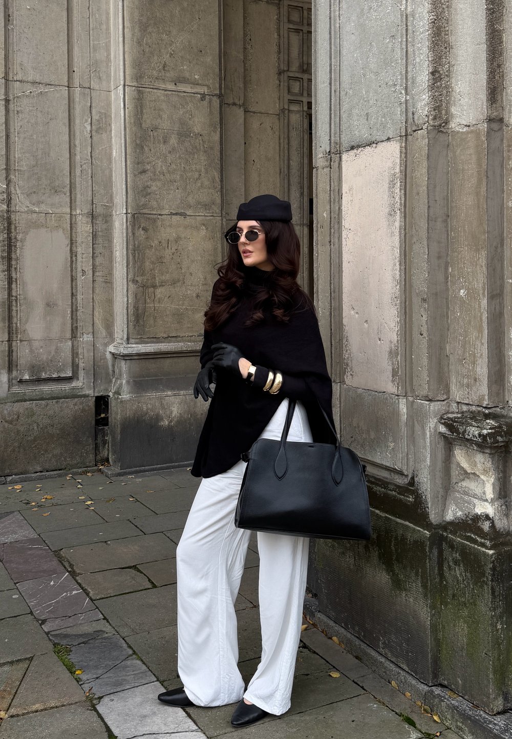 Poncho noir, pantalon large blanc, sac à main noir, chaussures noires à talons pointus, bracelets en or, lunettes de soleil rondes, avec un béret, appuyée contre un mur.