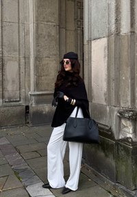 Poncho noir, pantalon large blanc, sac à main noir, chaussures noires à talons pointus, bracelets en or, lunettes de soleil rondes, avec un béret, appuyée contre un mur.