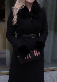 Manteau en laine noire avec col et poignets en fausse fourrure, ceinturé à la taille. Tient une pochette noire texturée. Tissu lisse, coupe ajustée.