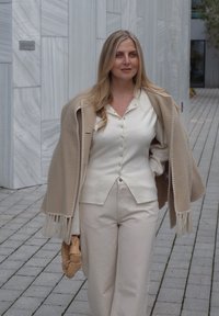 Bēša, ar bārkstīm apdarināta šalle pār baltu krekliņu ar pogām. Gaiši bēšas, plašas bikses. Rokās pinuma brūna soma. Fona teksturēta marmora siena.