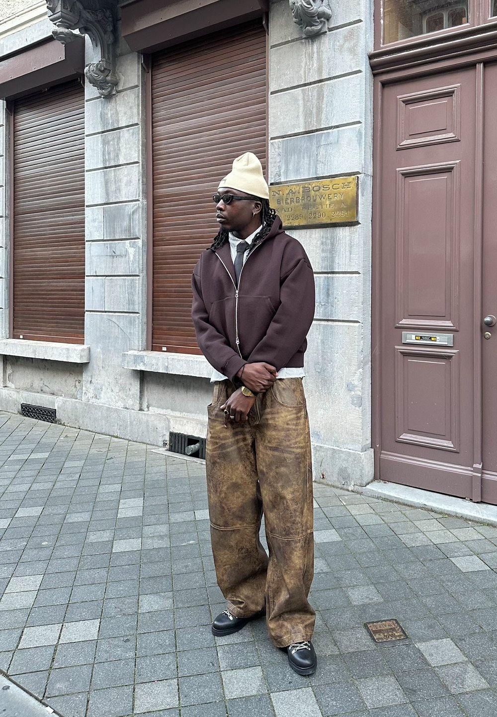 Brauner Reißverschluss-Hoodie, weißes Hemd mit Krawatte, braune strukturierte weite Hose, Sonnenbrille, beiger Beanie, steht auf gepflasterter Straße neben einer Steinmauer.