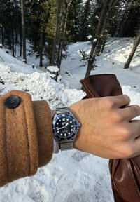 Πρόσωπο καρπού με ασημένιο ρολόι Timex και μανσέτα καφέ παλτού, κρατώντας καφέ δερμάτινη τσάντα, δάσος και δέντρα καλυμμένα με χιόνι στο φόντο.