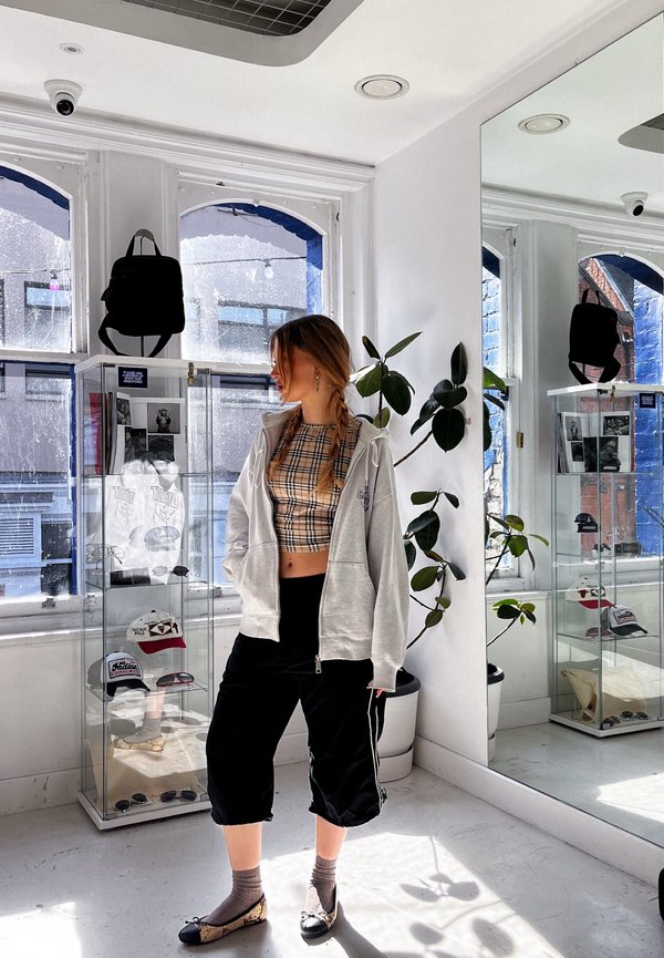 Tânără cu părul împletit stă într-o cameră luminată de soare purtând un crop top în carouri, hanorac gri, pantaloni negri și balerini cu model în apropierea vitrinelor.