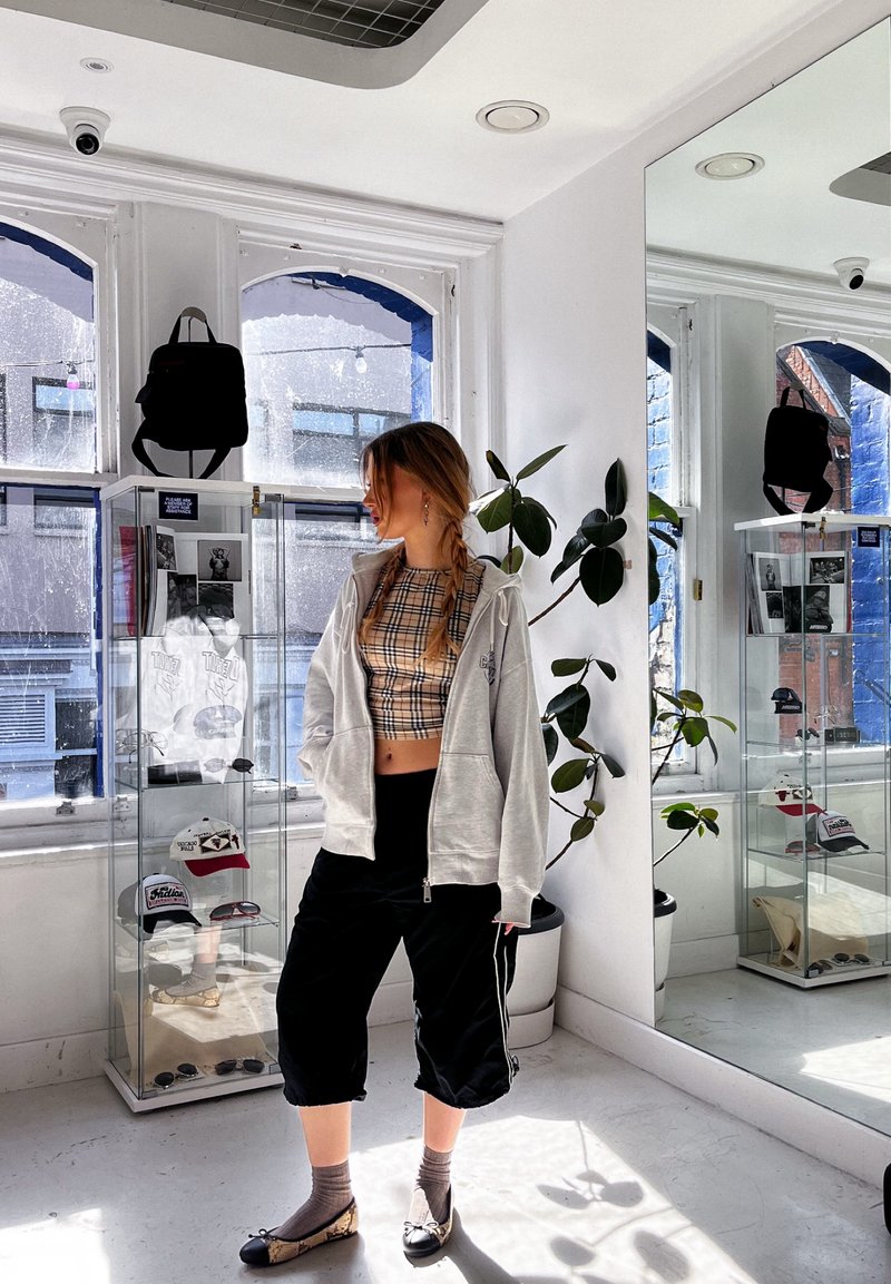 Jeune femme aux cheveux tressés se tient dans une pièce ensoleillée, portant un crop top à carreaux, un sweat à capuche gris, un pantalon noir et des ballerines à motifs près des vitrines.