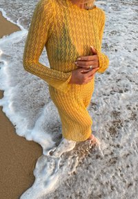 Personne debout pieds nus dans une écume de mer peu profonde sur le sable, portant une robe en dentelle jaune, les mains doucement jointes à la taille.