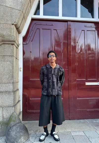 Camisa oversized negra con patrón y cuello, pantalones cortos negros, calcetines negros y zapatos bicolores, con un fondo de puerta roja.