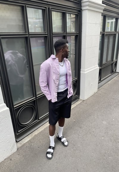 Camisa de botones rosa claro, camiseta blanca de tirantes, pantalones cortos negros, calcetas blancas, sandalias negras con detalles plateados, de pie contra una pared de vidrio.