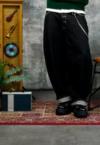 Personne portant un jean noir à jambes larges et des bottes noires debout sur un tapis rouge à motifs près d'une étagère en bois avec des instruments de musique et une radio vintage.