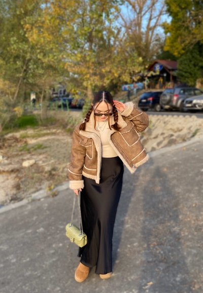 Chaqueta de borrego marrón con forro crema, camiseta beige, falda larga negra y botas beige. Sosteniendo un pequeño bolso verde. Entorno al aire libre.