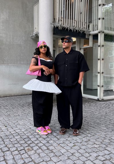 Camisa y pantalones holgados negros combinados con falda blanca con pliegues; bolso rosa y zapatos con acentos verdes; gafas de sol completan el look.