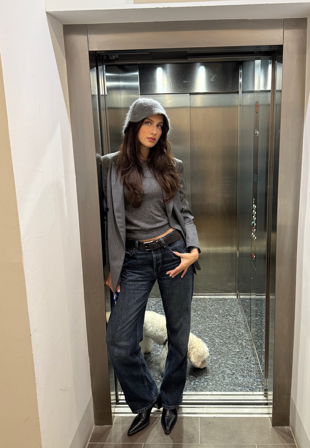 Graue Wollmütze, graues Oberteil, gestreifter Blazer, dunkle Jeans mit weitem Bein, schwarze spitze Absätze; Aufzugshintergrund mit grauen und metallischen Akzenten.